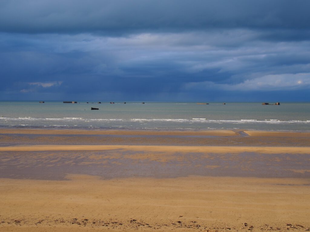 Asnelles Beach med en overskyet himmel, gyldne sandklitter og maritime rester, der er synlige i det fjerne.