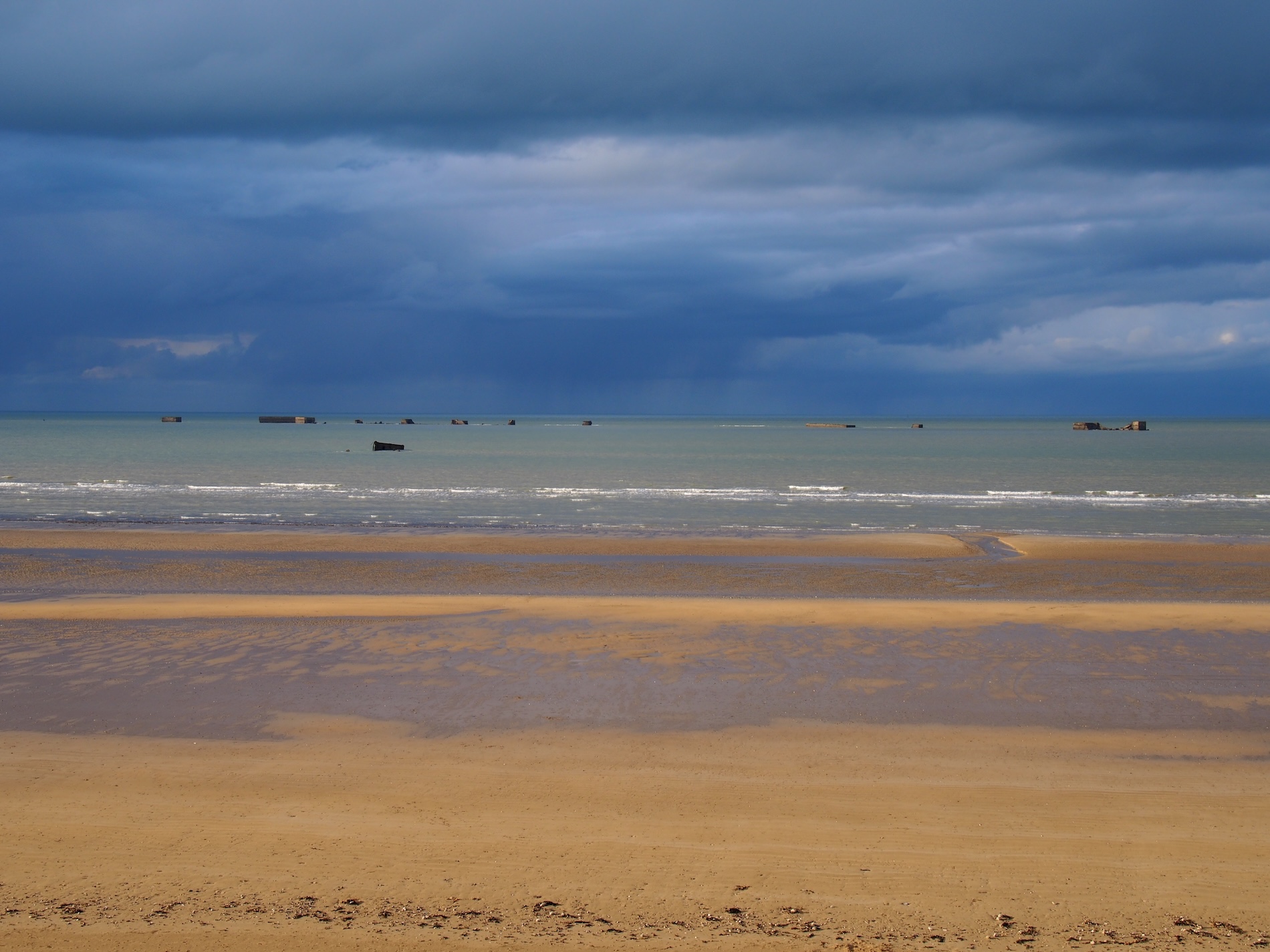 Asnelles Beach med en overskyet himmel, gyldne sandklitter og maritime rester, der er synlige i det fjerne.