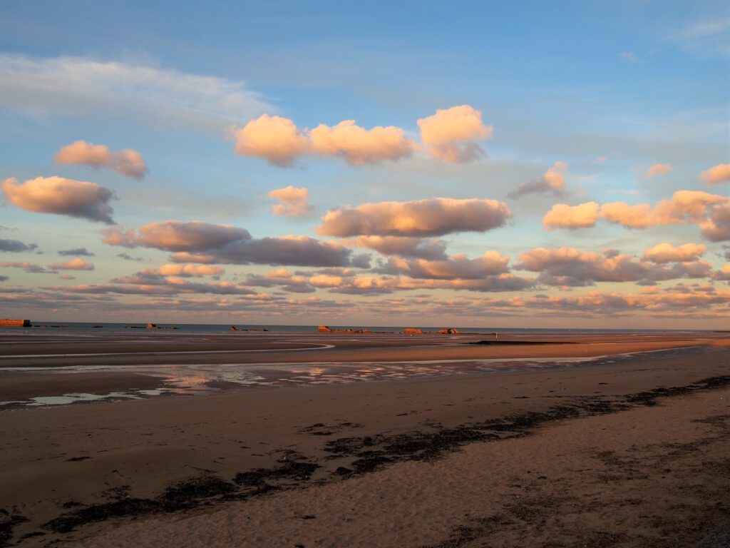Asnelles Beach ved solnedgang, med bløde skyer og rester på kysten, hvilket skaber en fredelig atmosfære.