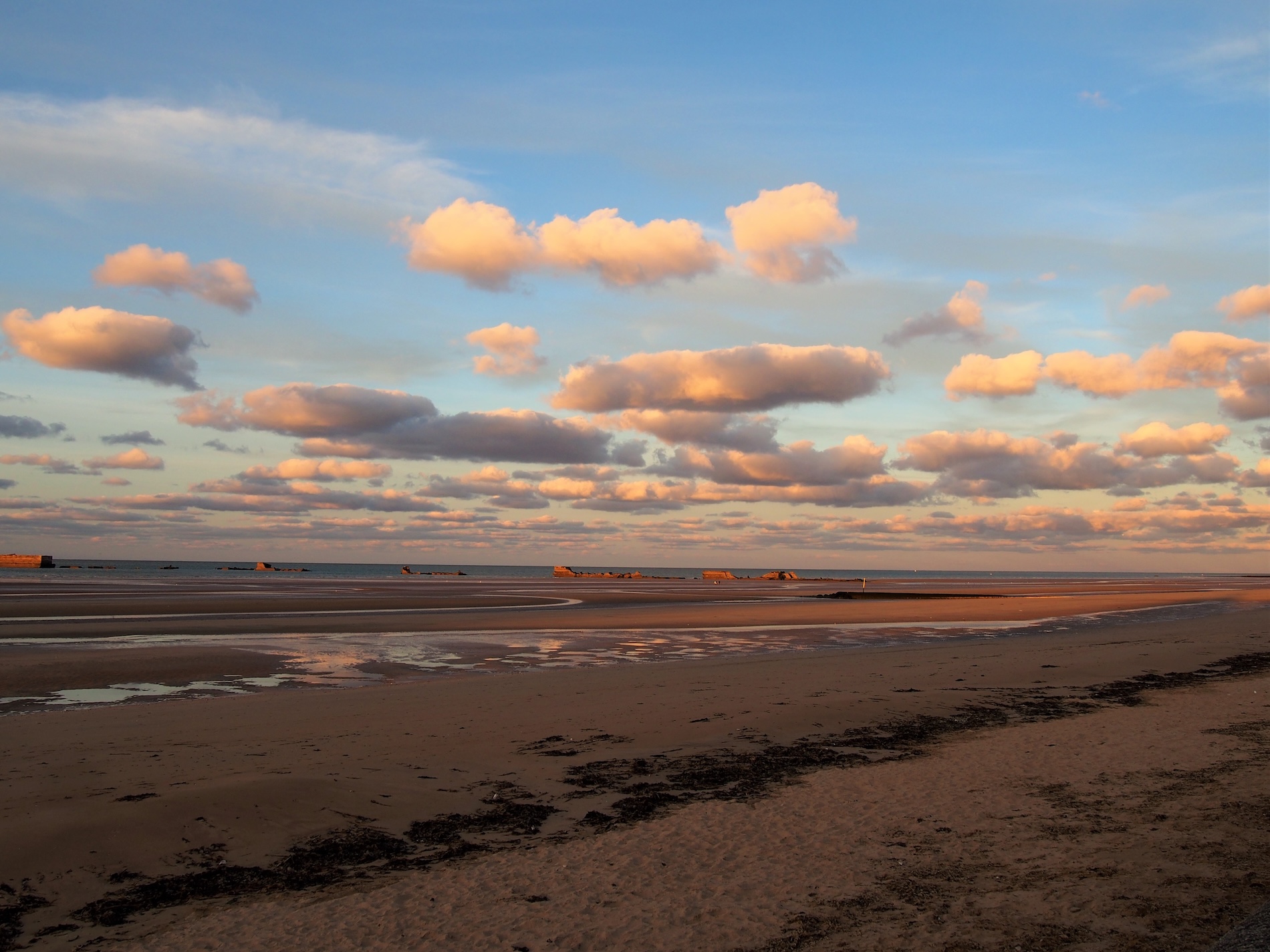 Asnelles Beach ved solnedgang, med bløde skyer og rester på kysten, hvilket skaber en fredelig atmosfære.