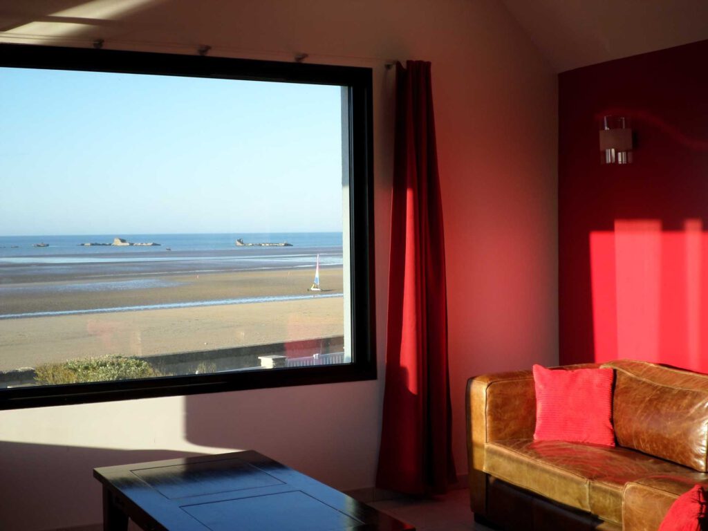 Indvendigt udsigt over et sommerhus i Asnelles, med en lædersofa og et vindue med udsigt over stranden.