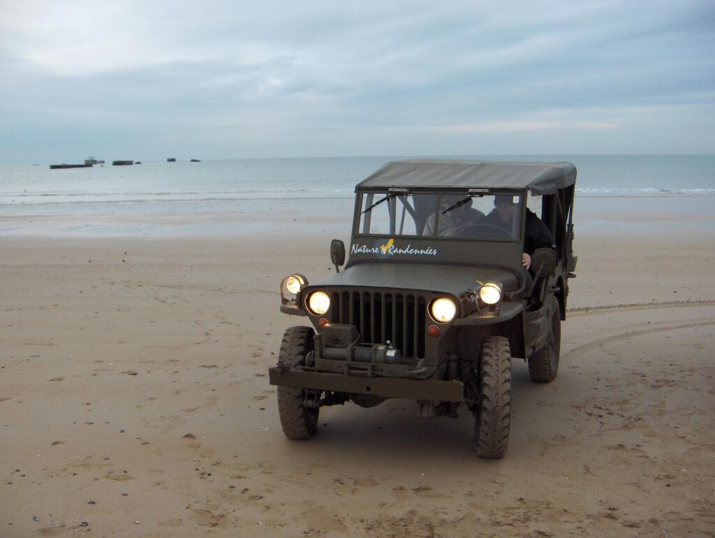 Et militært køretøj ruller på en normandisk strand, med havet i baggrunden og rester, der er synlige på vandoverfladen.
