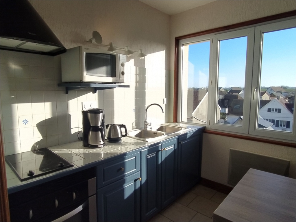 Lys køkken med blå bordplader, moderne udstyr og vindue, der tilbyder udsigt over landsbyen Asnelles.
