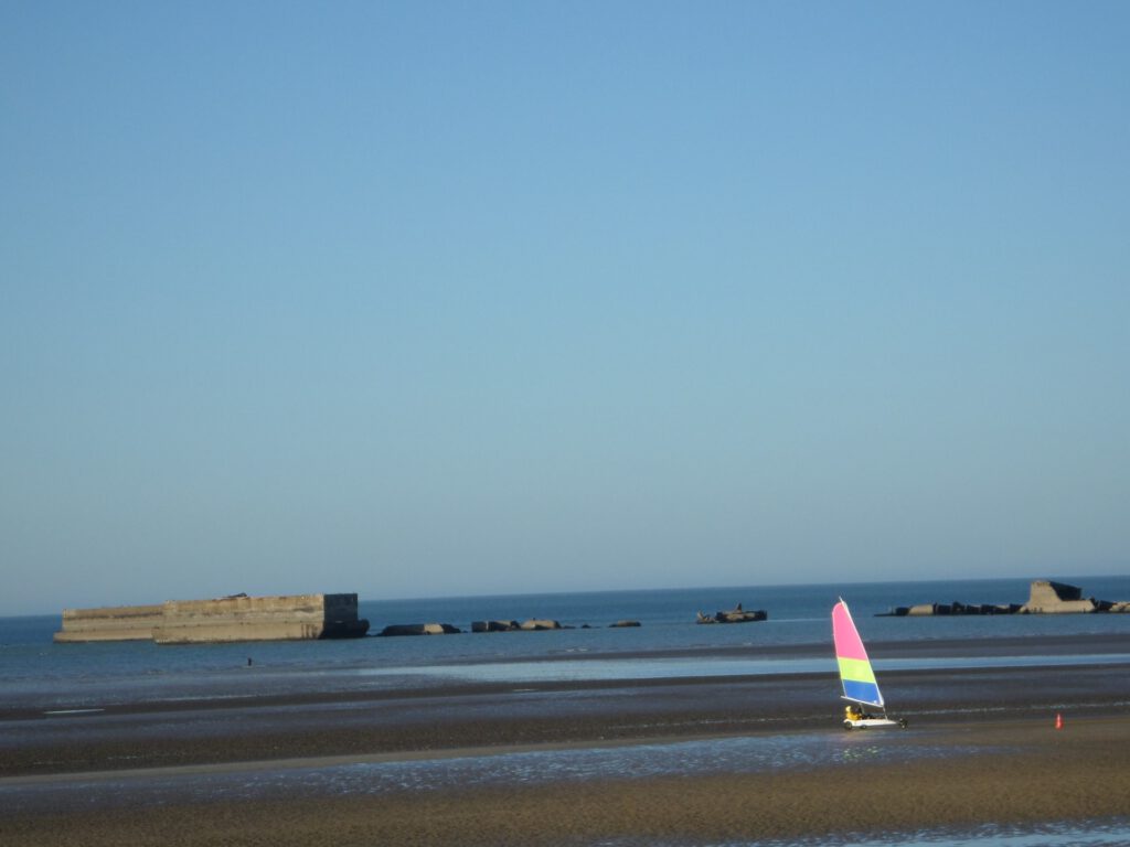 Farverigt nautisk køretøj på en Normandy strand med klipper og et roligt hav i baggrunden.