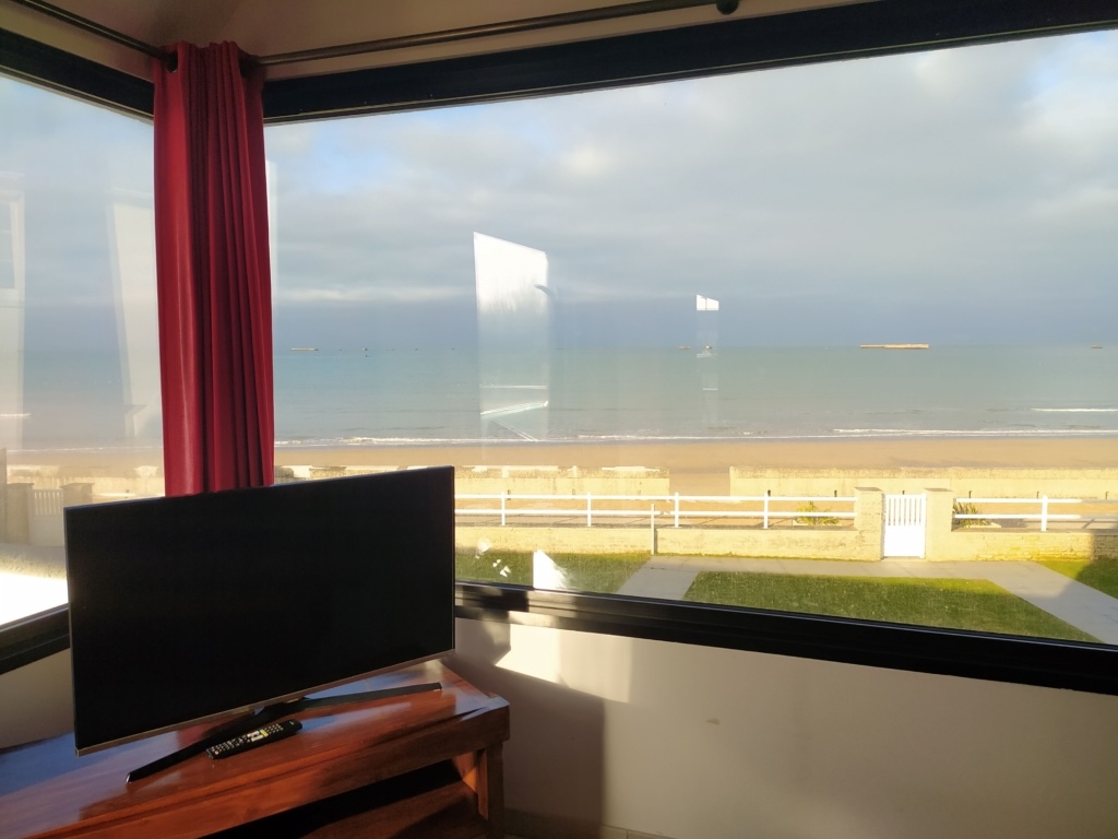 Panoramaudsigt over havet fra et sommerhus i Asnelles med et tv og røde gardiner.